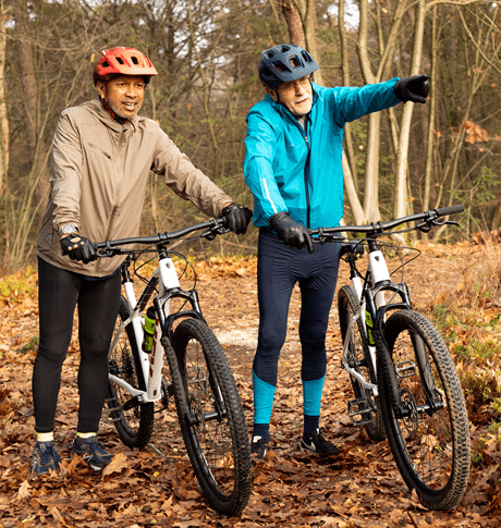 Twee mannen met mountainbikes in fietskleding in het bos staan stil naast hun fiets. Zij kijken naar iets in de verte en een van de mannen wijst ernaar.
