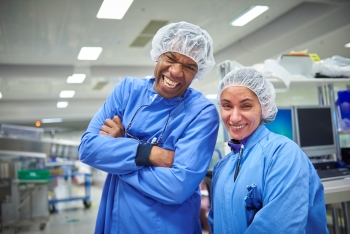 Een man en een vrouw in de productie kijken lachend in de camera. Zij hebben beschermende kleding aan – een haarnetje en een blauwe overjas.