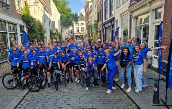 A group of around 30 cyclists are standing together in the Matchis – Amgen cycle outfits cheering and smiling. They hold a sign on the amount they have gathered to sponsor Matchis by cycling the Fietselfstedentocht.  
