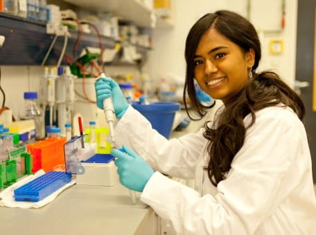 A young woman wearing a white lab coat stands in the laboratory with a pipette in her hand and looks into the camera, smiling.
