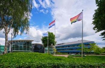 The Amgen building in Breda seen from the frontside of the building with the three flags one with an Amgen logo, the Dutch flag and a Pride flag. The photo also shows a lot of greenery, namely trees in the garden.