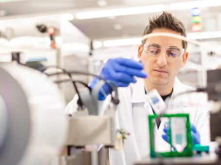 An Amgen lab technician and scientist wearing goggles and blue gloves looks intently at a vial he holds in his hands.