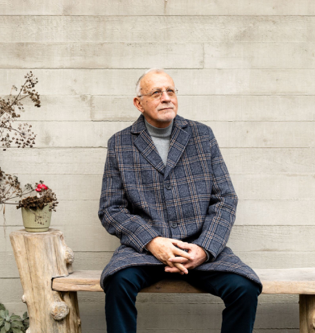 Een man zit op een houten bankje voor een grijze verweerde houten schuting. Hij staart in de verte. Naast hem staan verdroogde bloemen.